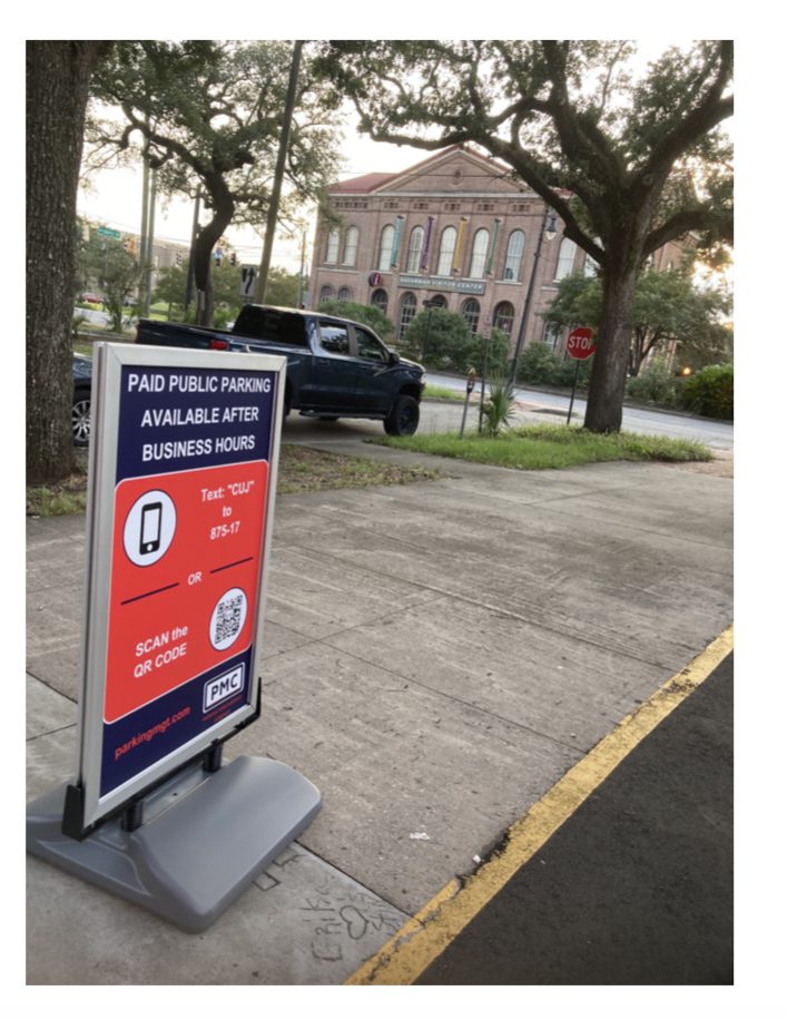 A sidewalk sign with the PMC logo in the bottom right corner reads “Paid public parking available after business hours” with instructions to text or scan a QR code. In the background are trees, a black truck, and a historic brick building.