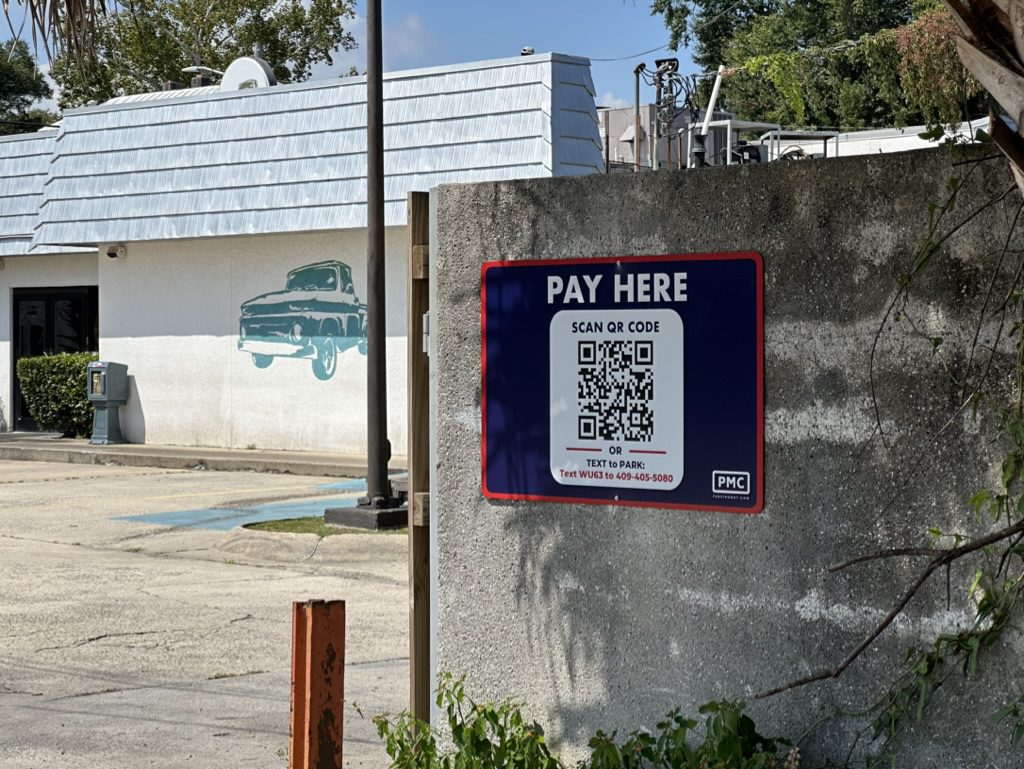 A "Pay Here" sign with a QR code and PMC logo is posted on a concrete wall in a Parking Management Company-owned parking lot. A white building with a vintage car mural is in the background.