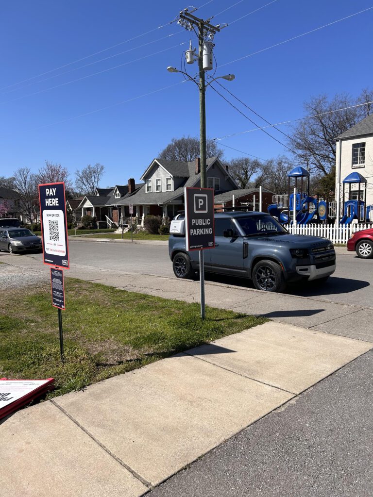 A gray SUV is parked on a street next to a public parking sign. Nearby, “Pay Here” parking signs with QR codes are visible. Houses, trees, and a playground are in the background under a clear blue sky.