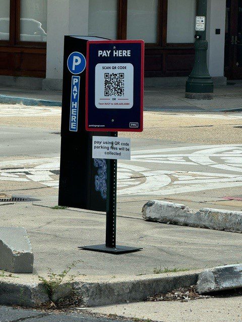 A "Pay Here" parking sign with a QR code stands near a curb and a parking meter on a city street. Instructions for scanning the code to pay fees are visible on the sign.