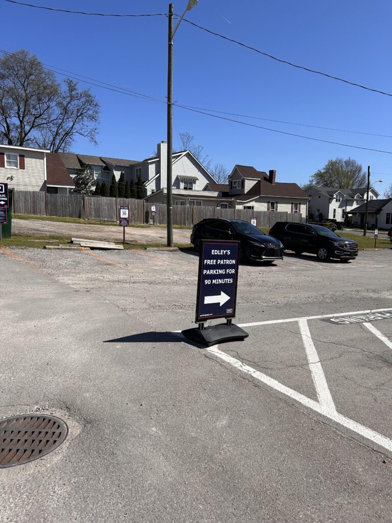 A sign in a parking lot reads "Edley's Free Patron Parking for 90 Minutes" with an arrow pointing right. Several parked cars and houses are visible in the background under a clear, blue sky.