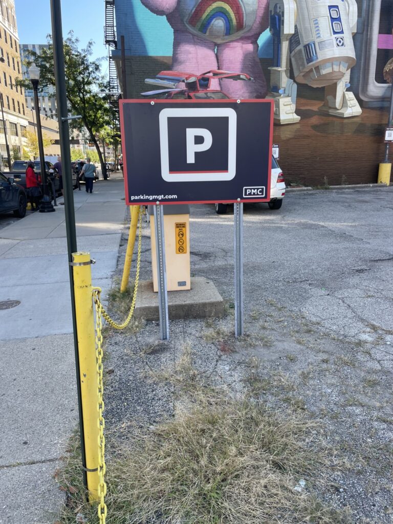 A parking sign stands in a lot near a yellow post and chain. In the background, there’s a colorful mural on a building wall featuring a robot and a rainbow design. Some people and parked cars are visible to the left.