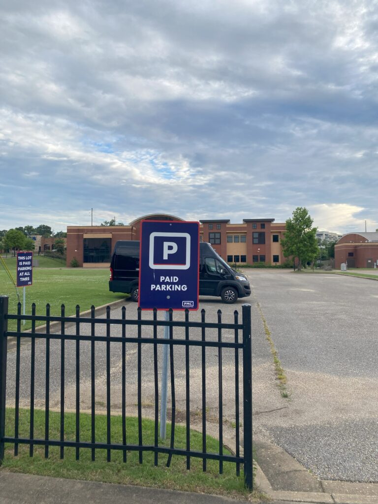 A parking lot with a "Paid Parking" sign near a black gate, a black van parked inside, and a brick building in the background under a cloudy sky.