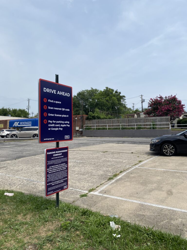 A parking lot with a car parked and a large sign displaying instructions for paying for parking using a QR code, credit card, Apple Pay, or Google Pay. A store and trees are visible in the background under a cloudy sky.