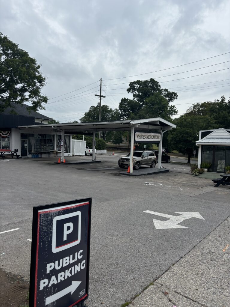 A sign in the foreground reads "Public Parking" with an arrow pointing left. In the background is a small building labeled "White's Mercantile" and a parked car under a carport on a cloudy day.