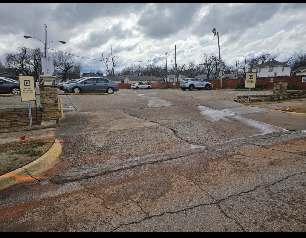 A mostly empty parking lot on a cloudy day with a few cars parked. Two reserved parking signs are visible, and there are cracks and patches on the pavement. Leafless trees and utility poles line the background.