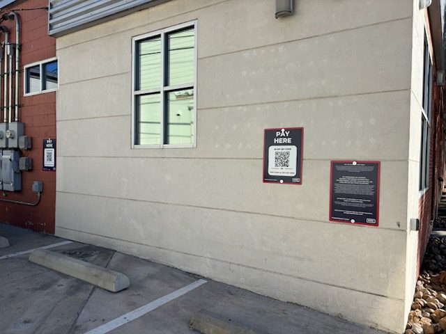 A parking lot with empty parking spaces beside a beige building. Signs on the wall include a "Pay Here" sign with a QR code and another sign with parking instructions. Utility meters are visible on the left.