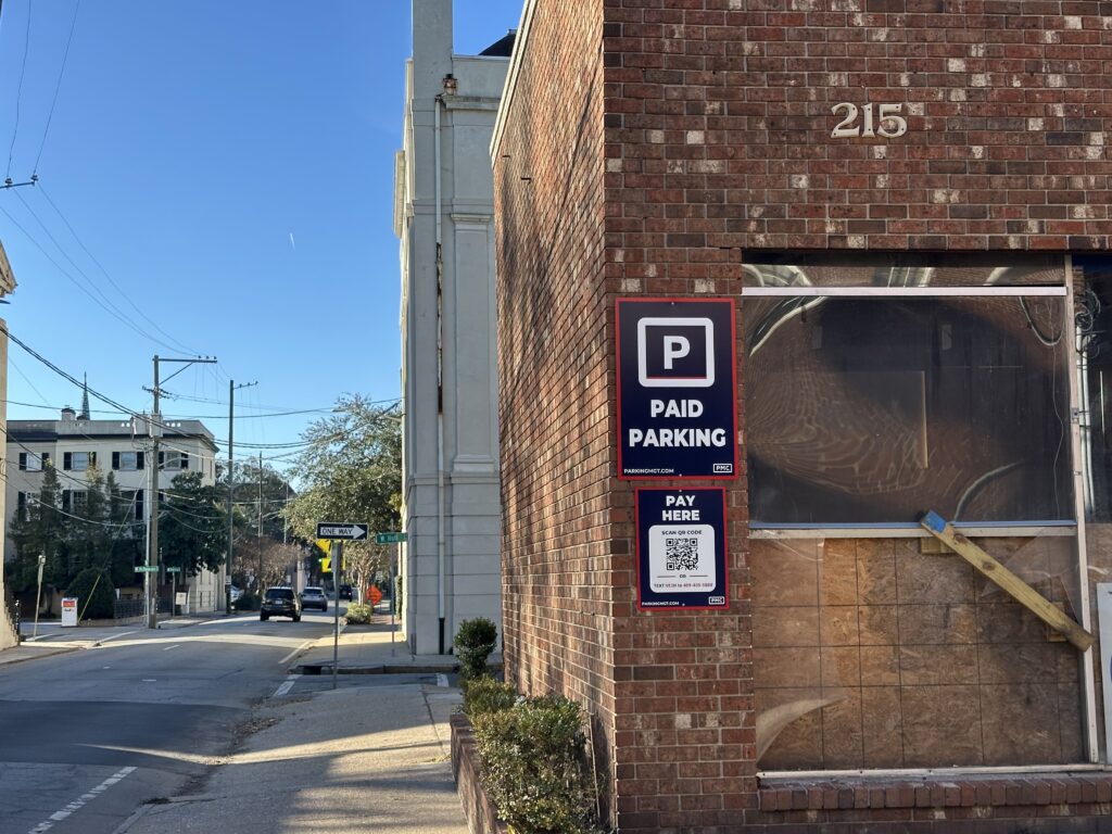 A brick building with a PMC “Paid Parking” sign and a QR code for payment on its wall, located on a quiet city street with sidewalks, trees, and distant buildings under a clear blue sky.