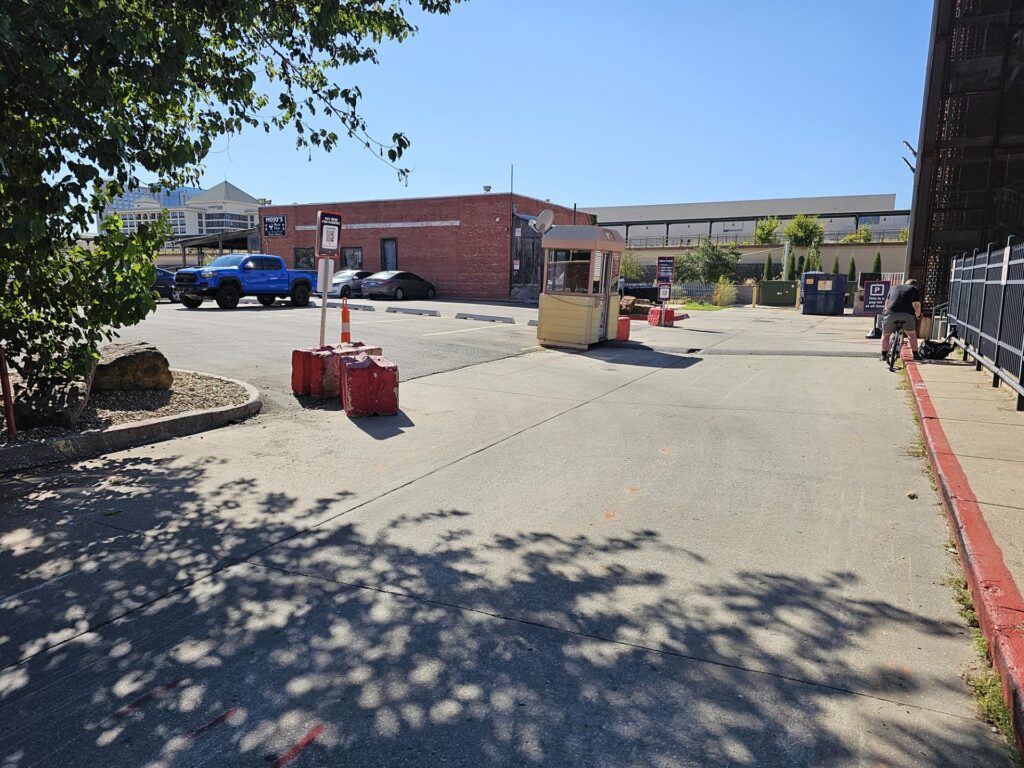 A PMC-owned paved parking lot with a small yellow booth, red barriers, and a few parked cars under a clear blue sky.