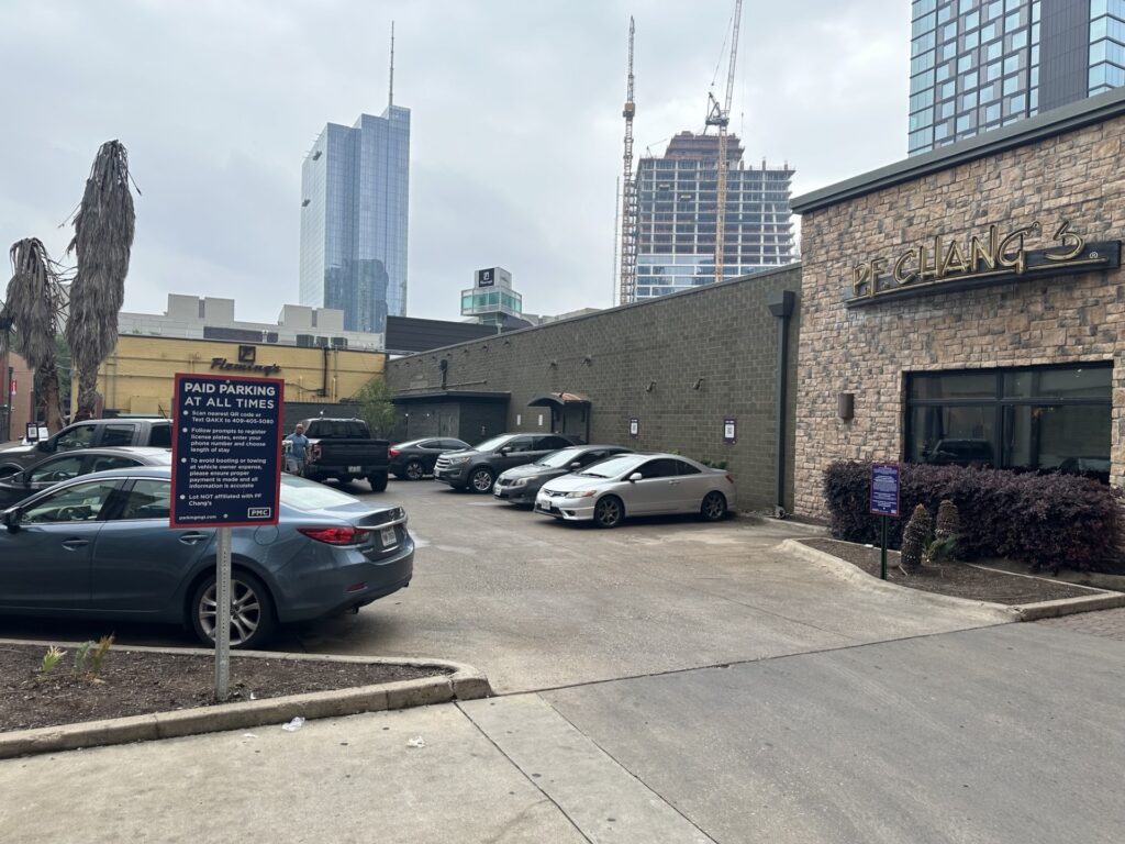 A small parking lot with several parked cars beside a stone building labeled "P.F. Chang's." A sign in the lot reads "PAID PARKING AT ALL TIMES." Tall buildings and construction cranes are visible in the background.