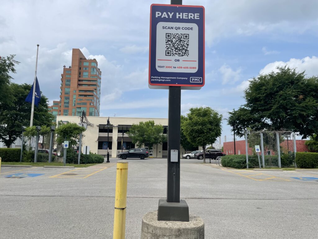 A parking lot with a tall sign reading "PAY HERE" featuring a QR code for payment. In the background are trees, parked cars, accessible parking spaces, and a multi-story brick building under a partly cloudy sky.