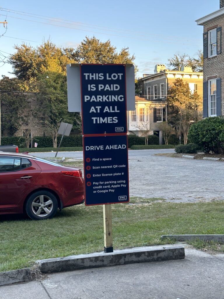 A PMC sign in front of a parking lot reads, “This lot is paid parking at all times.” Instructions below explain how to pay via QR code, enter the license plate, and use credit card, Apple Pay, or Google Pay.
