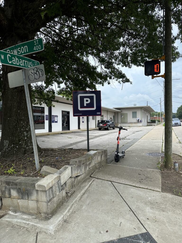 A sidewalk scene with a parking sign, street signs for Dawson St and W Cabarrus St, a parked scooter, and a countdown crossing signal displaying 10. Buildings and cars are visible in the background.