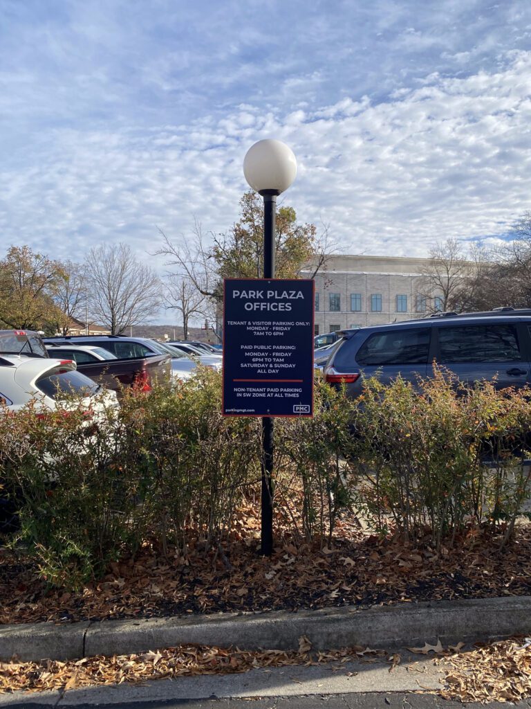 A black sign for Park Plaza Offices stands on a lamppost in a parking lot, surrounded by shrubs and parked cars. Trees and a large building are visible in the background under a partly cloudy sky.