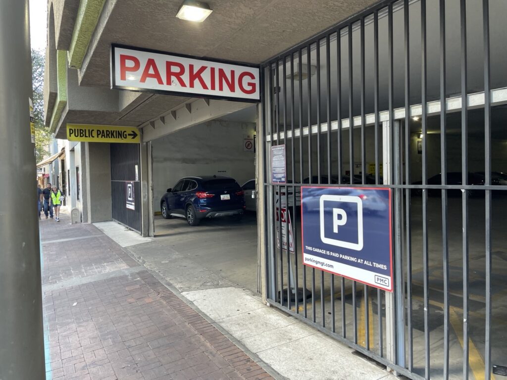 A parking garage entrance with a red and white "PARKING" sign above, a yellow "PUBLIC PARKING" sign above to the left, and a large blue PMC parking sign on a metal gate to the right indicating parking hours and website information. Two cars are visible inside the garage.