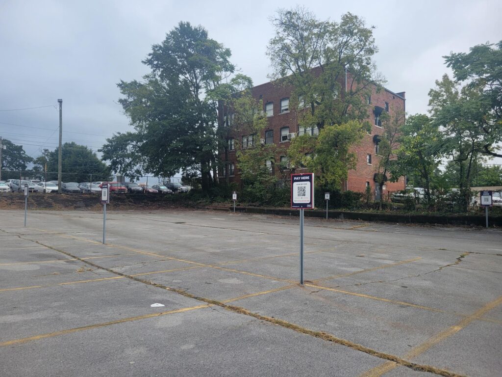 An empty parking lot with several reserved parking signs, yellow parking lines, and a few trees. In the background, there is a red brick apartment building and overcast sky.