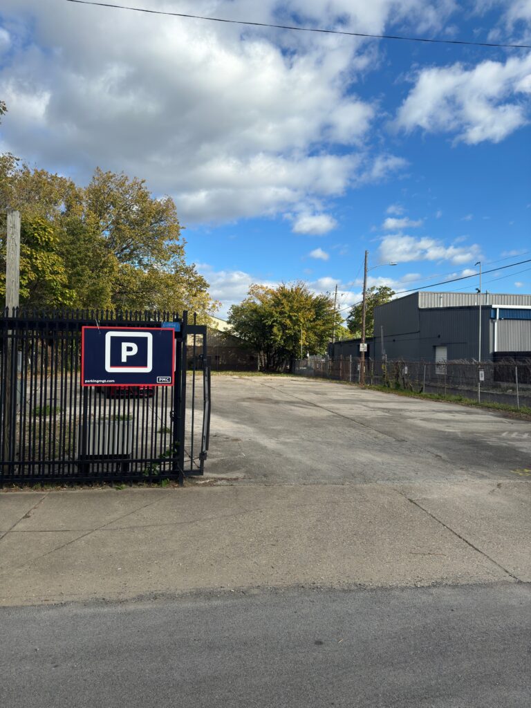A mostly empty parking lot is visible behind a black metal gate with a large "P" parking sign. Trees with green and yellow leaves and industrial buildings are in the background under a partly cloudy sky.