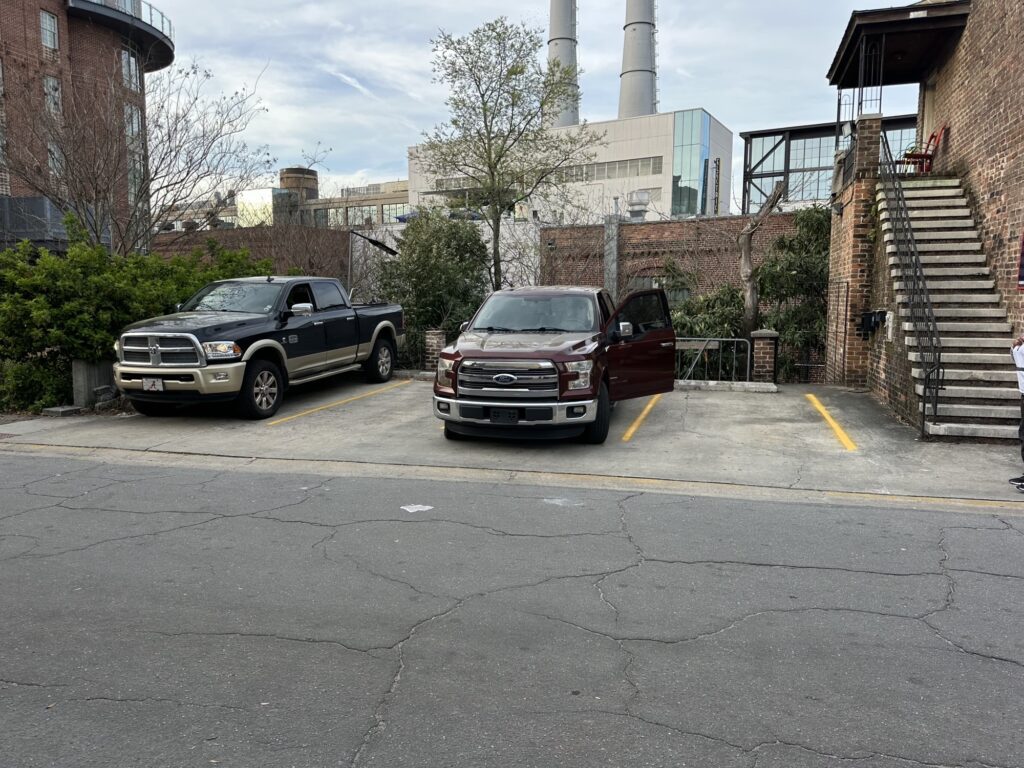 Two pickup trucks are parked in adjacent parking spots next to a brick building with an outdoor staircase. One truck has its driver's door open. Industrial buildings and trees are visible in the background.