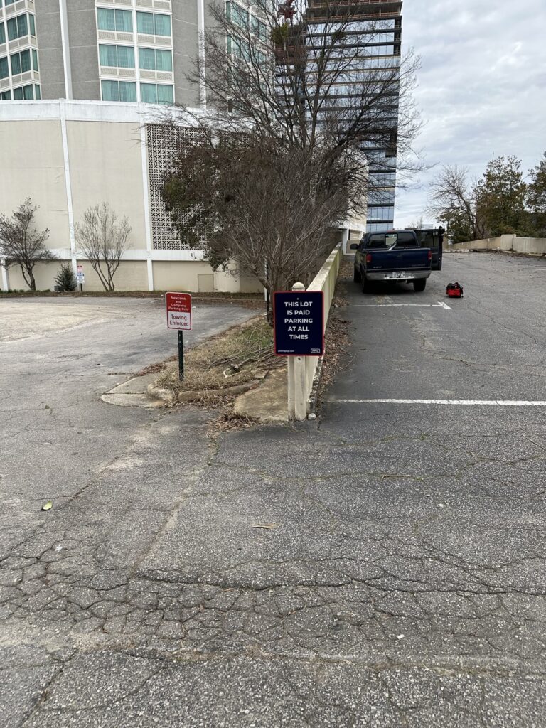 A PMC-owned parking lot with a black pickup truck parked near a beige building. Signs by a low wall say “This lot is reserved parking—violators will be towed.” Some trees and cloudy sky are visible in the background.