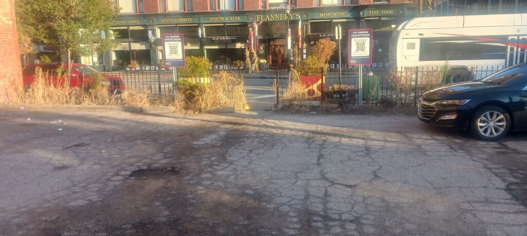A sunlit parking lot with cracked pavement, dry grass, two parked cars, and businesses, including Flannery's Pub, across the street. Two signs with QR codes are visible by a fence.