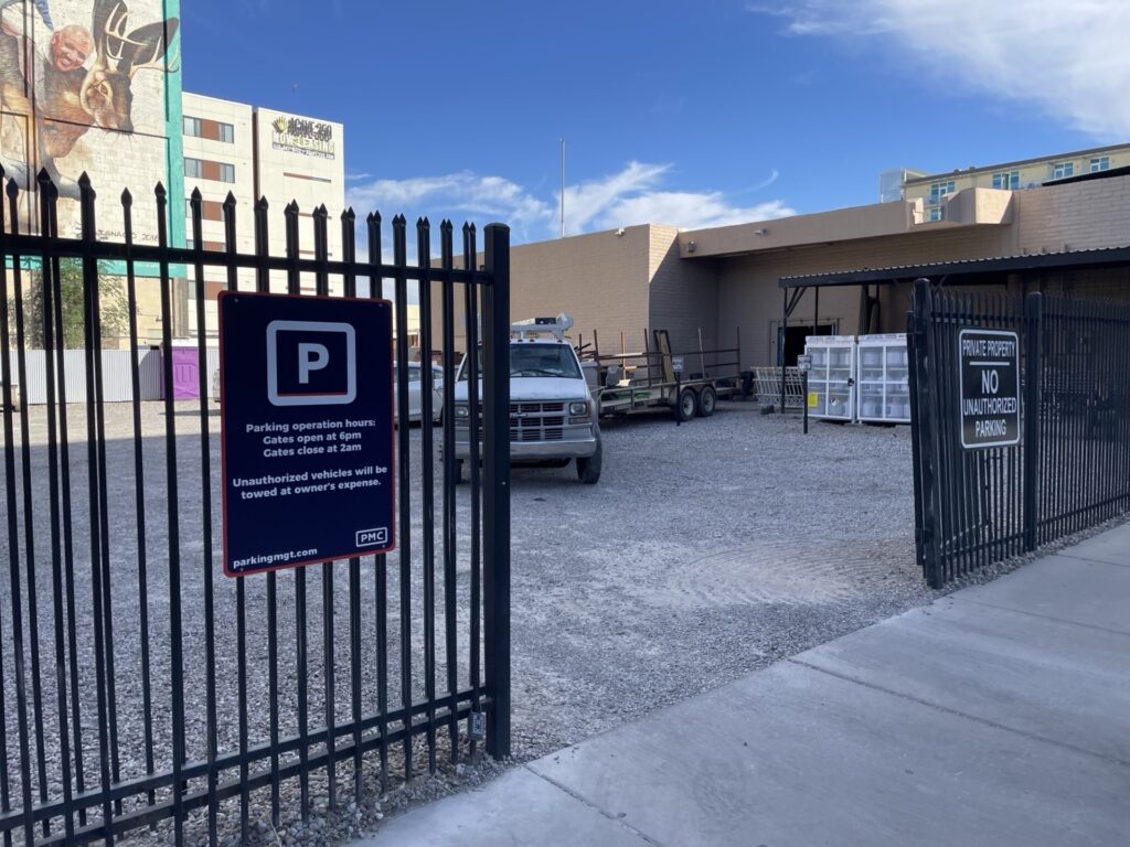 A fenced parking lot with a PMC sign showing parking operation hours and rules. Inside, a white truck with a trailer is parked near a building, and several white crates are stacked nearby. Blue sky and nearby buildings are visible.