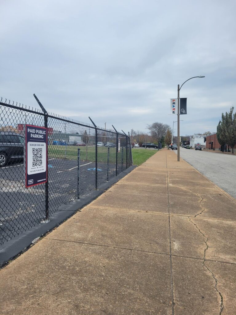 A sidewalk runs alongside a fenced Parking Management Company parking lot on a cloudy day. A PMC sign with a QR code for payment is attached to the fence with trees, cars, and buildings in the background.