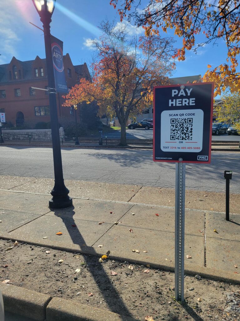 A PMC "Pay Here" sign with a QR code stands on a sidewalk under a streetlamp. Autumn trees and a brick building are visible in the background on a sunny day.