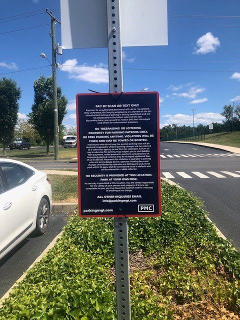 A parking sign on a post in a parking lot with rules and payment instructions. A white car is parked nearby, and trees and a road are visible in the background under a sunny sky.