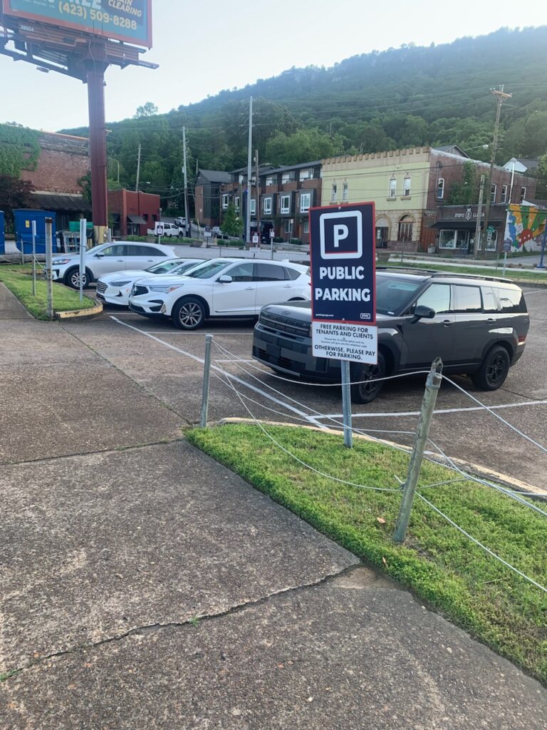 A public parking lot with several parked cars, a prominent “Public Parking” sign, and a row of small buildings in the background, set at the base of a green, tree-covered hill.