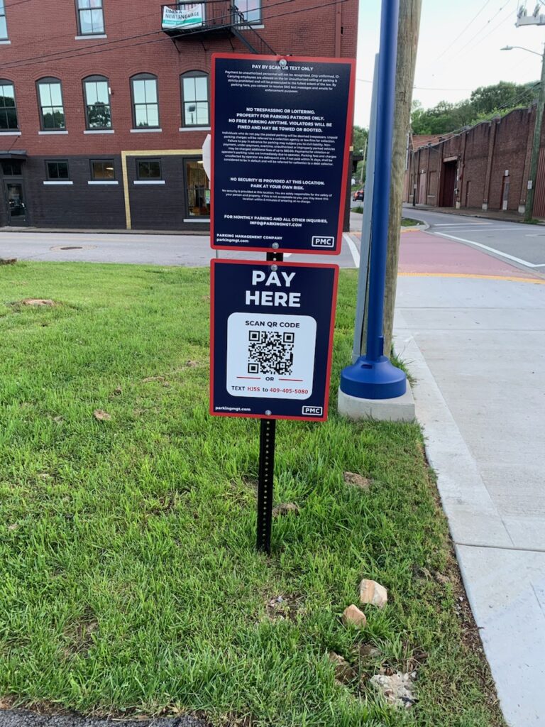 A sign on grass near a street displays instructions to "PAY HERE" with a large QR code for mobile payment and a phone number for assistance; brick buildings and a sidewalk are visible in the background.