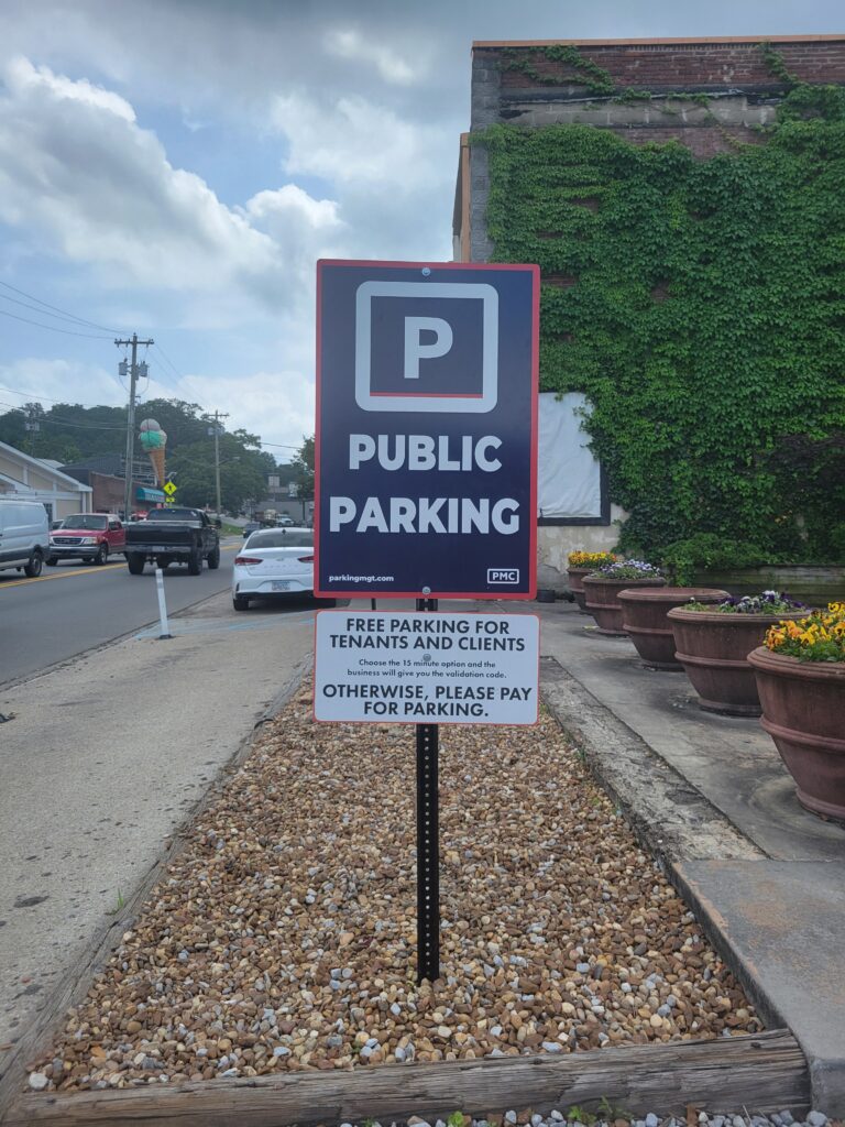 A black and white sign reads "Public Parking"; a smaller sign below says "Free parking for tenants and clients, otherwise please pay for parking." The sign is by a street, with cars and buildings visible under a cloudy sky.