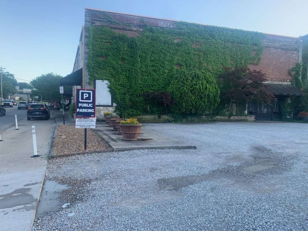 A gravel public parking lot next to a brick building covered in green ivy, with a "Public Parking" sign and large planters along the side. Several cars are parked along the street in the background.