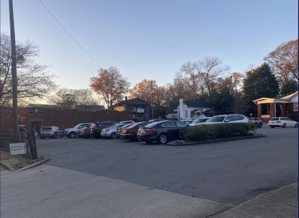 A small parking lot filled with cars is surrounded by trees with autumn leaves, residential houses, and a brick building under a clear evening sky.