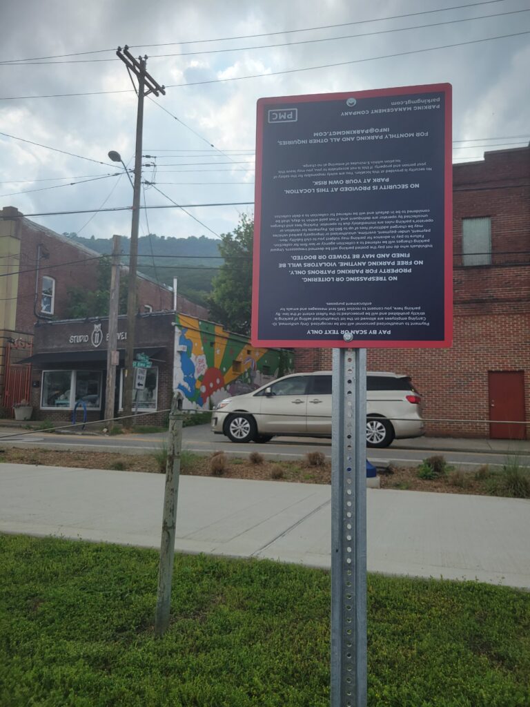 A street scene with a red and black sign mounted on a pole in the foreground. The sign is upside down. Behind it are a sidewalk, a white minivan, a mural on a building, and hills under a cloudy sky.