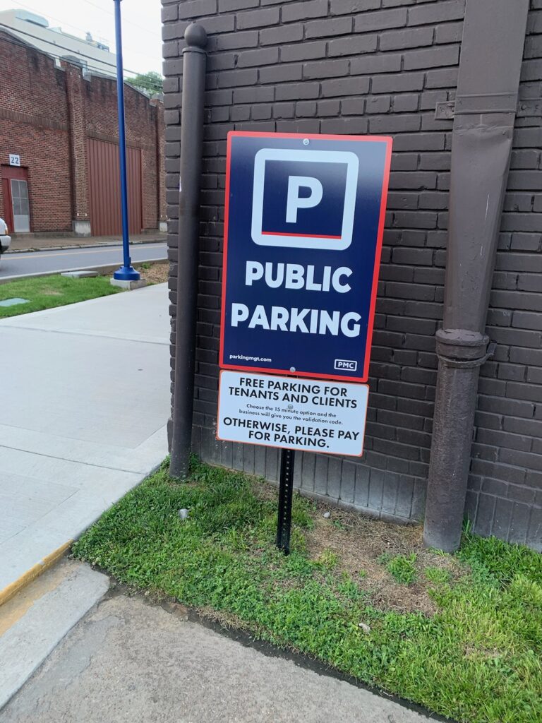 A sign next to a brick wall reads "Public Parking." Below, it says "Free parking for tenants and clients. Otherwise, please pay for parking." The sign is next to a sidewalk and grassy area.