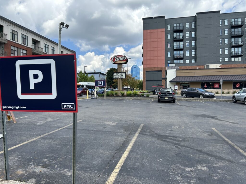A mostly empty parking lot with a large blue "P" parking sign in the foreground, several cars parked, a Raising Cane's restaurant, and a multi-story building in the background under a partly cloudy sky.