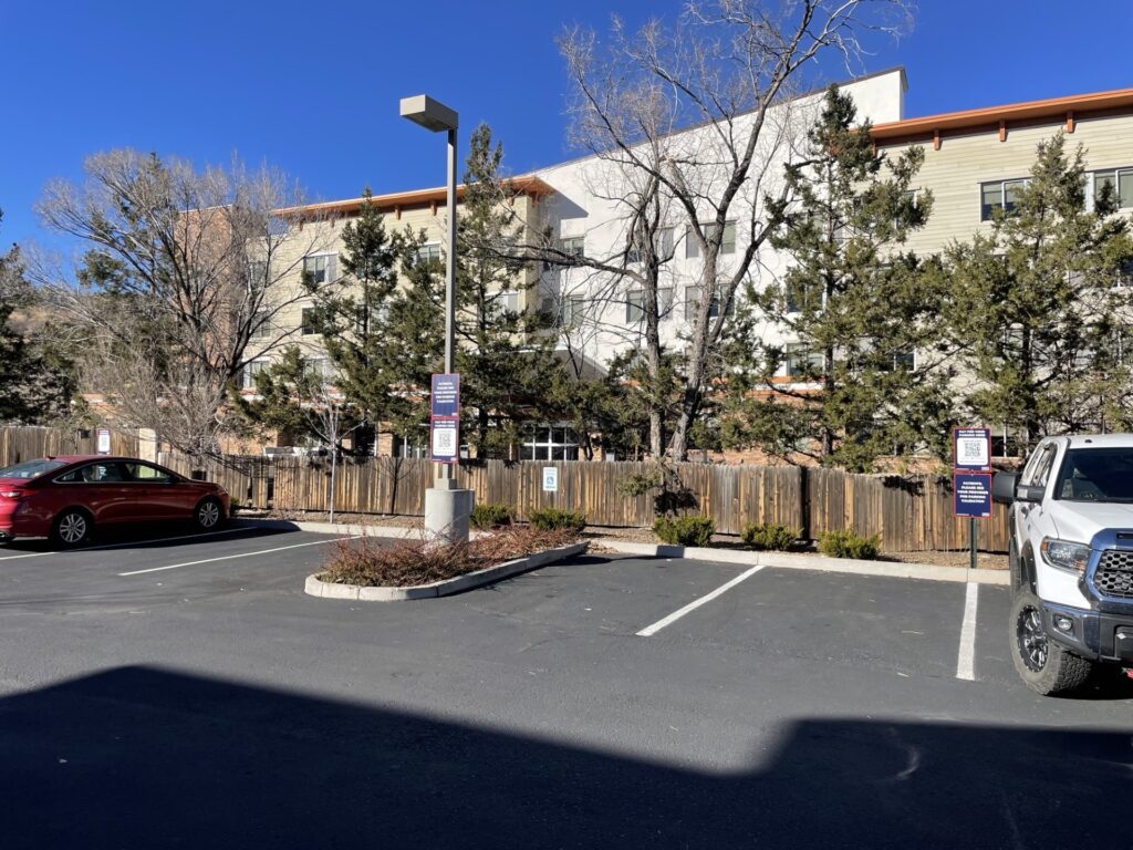 A small parking lot with a few parked cars, including a red sedan and a white truck, in front of a fenced area with trees and a multi-story building under a clear blue sky.