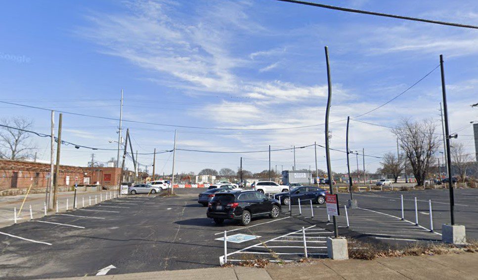 A mostly empty parking lot with a few cars parked, surrounded by power lines and a few bare trees, under a blue sky with scattered clouds. Some old buildings are visible in the background.