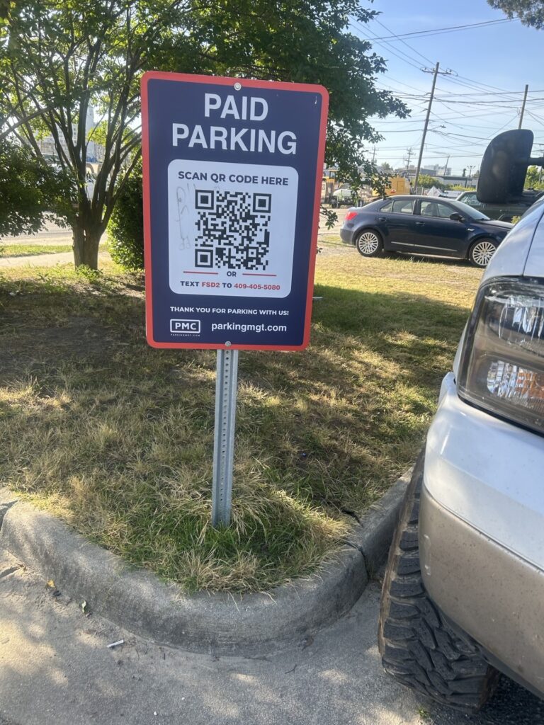 PMC sign in a parking lot reads “PAID PARKING.” It displays a QR code for payment and instructions to text a number to pay. The sign is on grass near a curb, with cars and trees in the background.