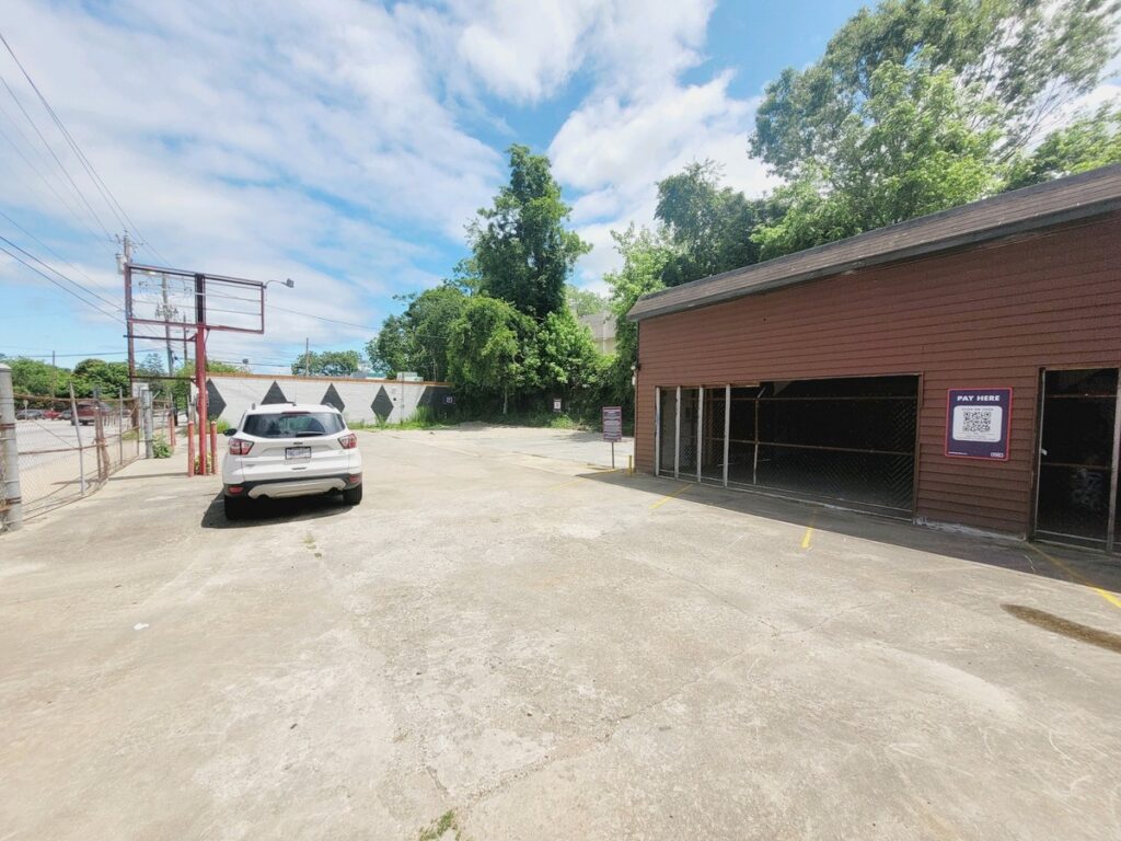 A mostly empty parking lot with a single white SUV parked on the left, next to a brown building with an open garage and a pay kiosk sign. Trees and a cloudy sky are visible in the background.