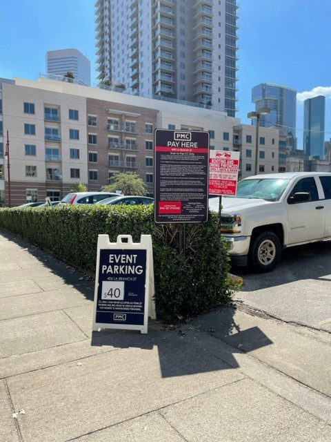 A sidewalk in a city with signs for PMC event parking, including a sandwich board showing a $40 parking fee. Cars are parked nearby and tall buildings are visible in the background under a clear, sunny sky.