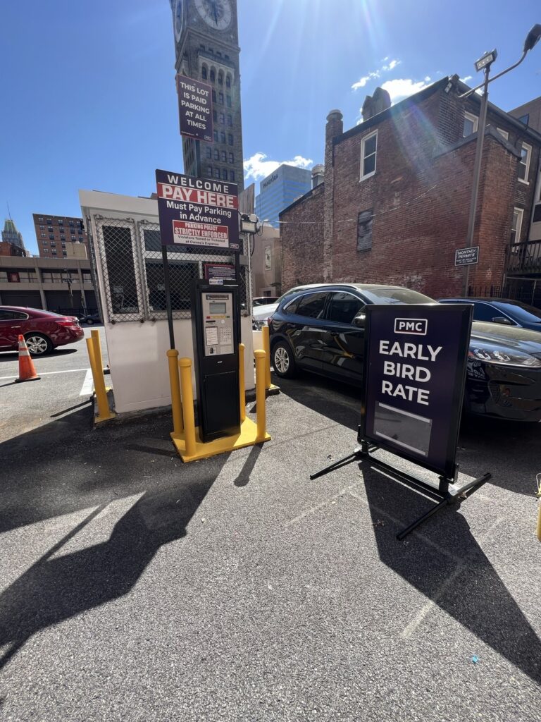 A parking lot entrance with a pay station, signs reading "WELCOME PAY HERE" and "PMC EARLY BIRD RATE," two parked cars, and a clock tower in the background under a sunny sky.