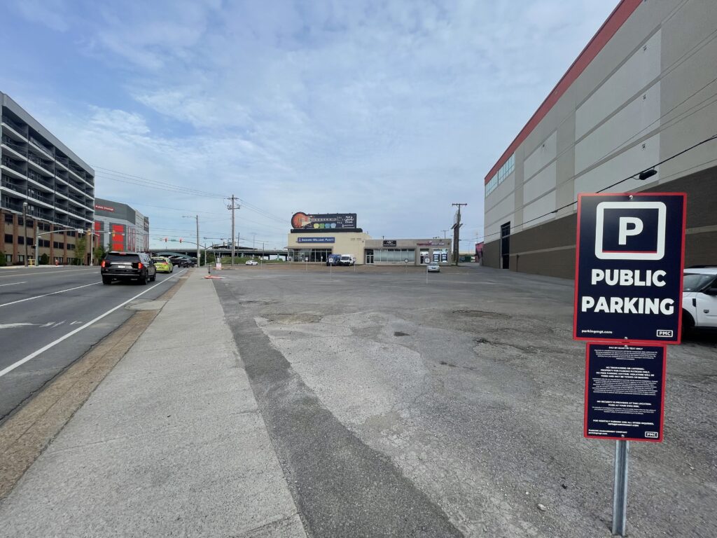 A mostly empty public parking lot next to a wide street, with a sign in the foreground that reads "Public Parking." Buildings and cars are visible in the background under a partly cloudy sky.