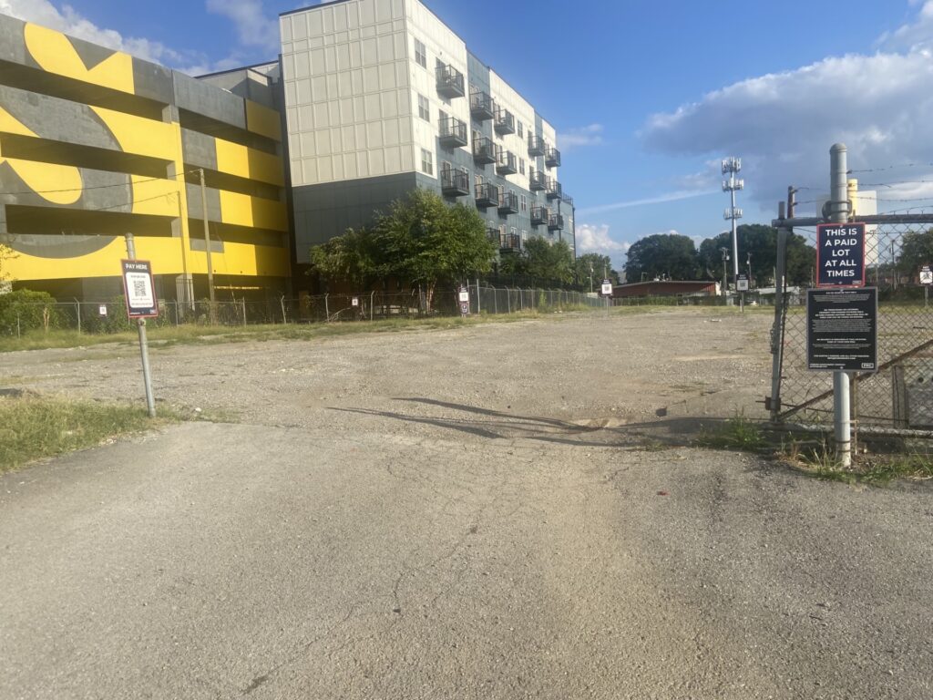 A mostly empty gravel parking lot surrounded by chain-link fencing, with signs on the fence. There’s a modern building with a bold yellow and black mural in the background under a partly cloudy sky.