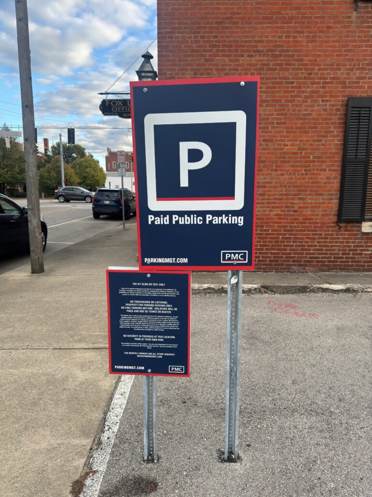 A sign in a parking lot reads "Paid Public Parking" with a large "P" symbol, posted on metal poles beside a brick building. The sky is partly cloudy and cars are parked nearby.