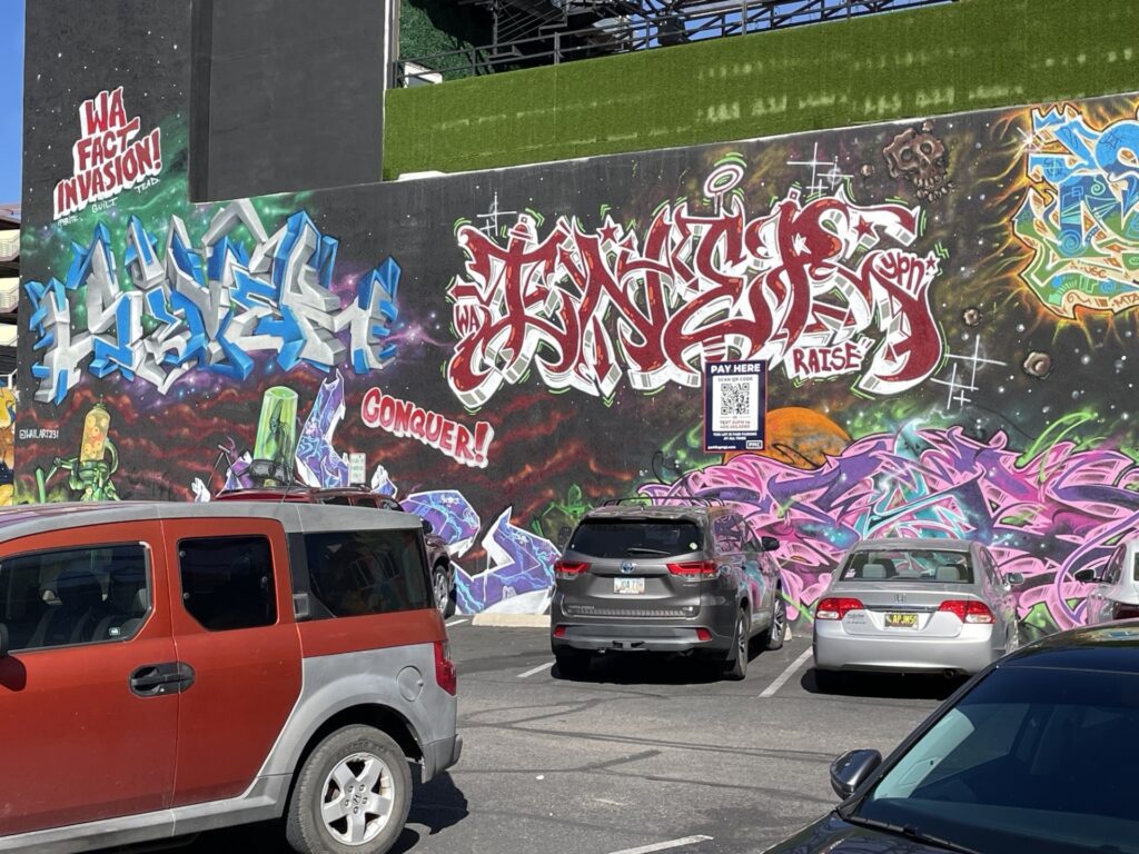 Three cars are parked in front of a colorful graffiti wall featuring a PMC sign with a QR code indicating to "Pay Here".