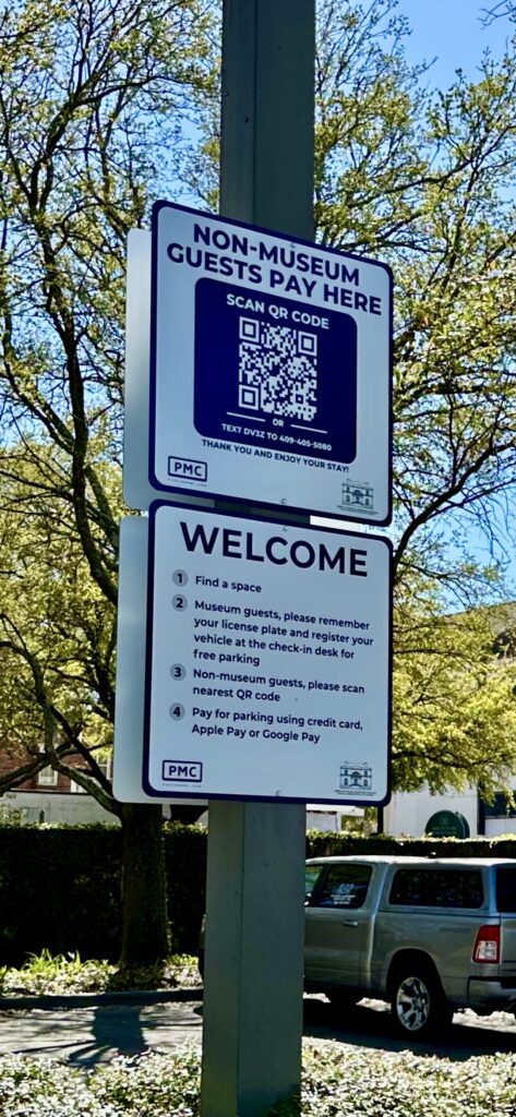 PMC parking sign on a street pole reads “Non-museum guests pay here” with a QR code and payment instructions. Below, it explains procedures for museum and non-museum guests, with trees and parked cars in the background.