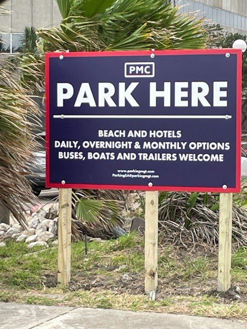 A sign reads: "PMC PARK HERE. Beach and hotels. Daily, overnight & monthly options. Buses, boats and trailers welcome." The sign is posted on grass near palm trees and a sidewalk.