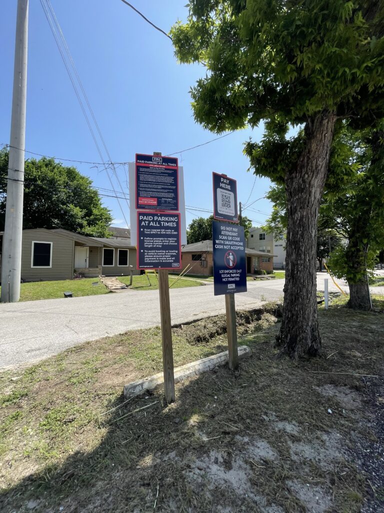 Two parking signs on wooden posts stand on a patch of grass beside a street, with houses and trees in the background under a clear blue sky. One sign displays a QR code.