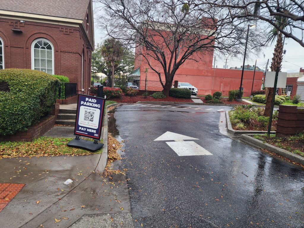 A wet parking lot with a white arrow painted on the ground. A sign on the left reads "Paid Parking" with a QR code and instructions to pay. Buildings, bushes, and a tree are in the background on a cloudy day.